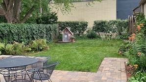 Eine Terrasse mit Tisch und Stühlen, eine kleine Gartenhütte und ein Pflasterweg, der zu einer Rasenfläche mit einem Baum und einem Zaun im Hintergrund führt.
