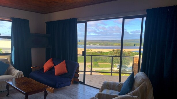 A spacious living room with a large window offering a view of a beach and ocean. There is a sofa with cushions and a wooden coffee table. - Spectacular Views of the bay -Bay View Witsand (Witsand)