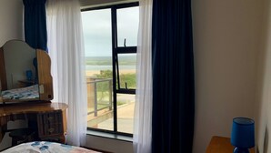 A bedroom with a bed, a dresser, a mirror, a window with curtains, and a view of a beach. - Spectacular Views of the bay -Bay View Witsand (Witsand)