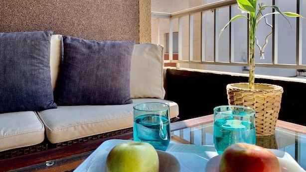 Un balcón con un sofá, dos cojines, una mesa de vidrio con dos vasos de agua, una planta en una cesta tejida y dos manzanas.