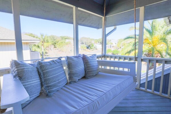 A covered porch with a white cushioned bench, a small table, and a view of palm trees and a beach.