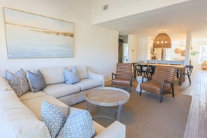 A living room with a sofa, patterned cushions, a coffee table, and a dining area with chairs and a chandelier.