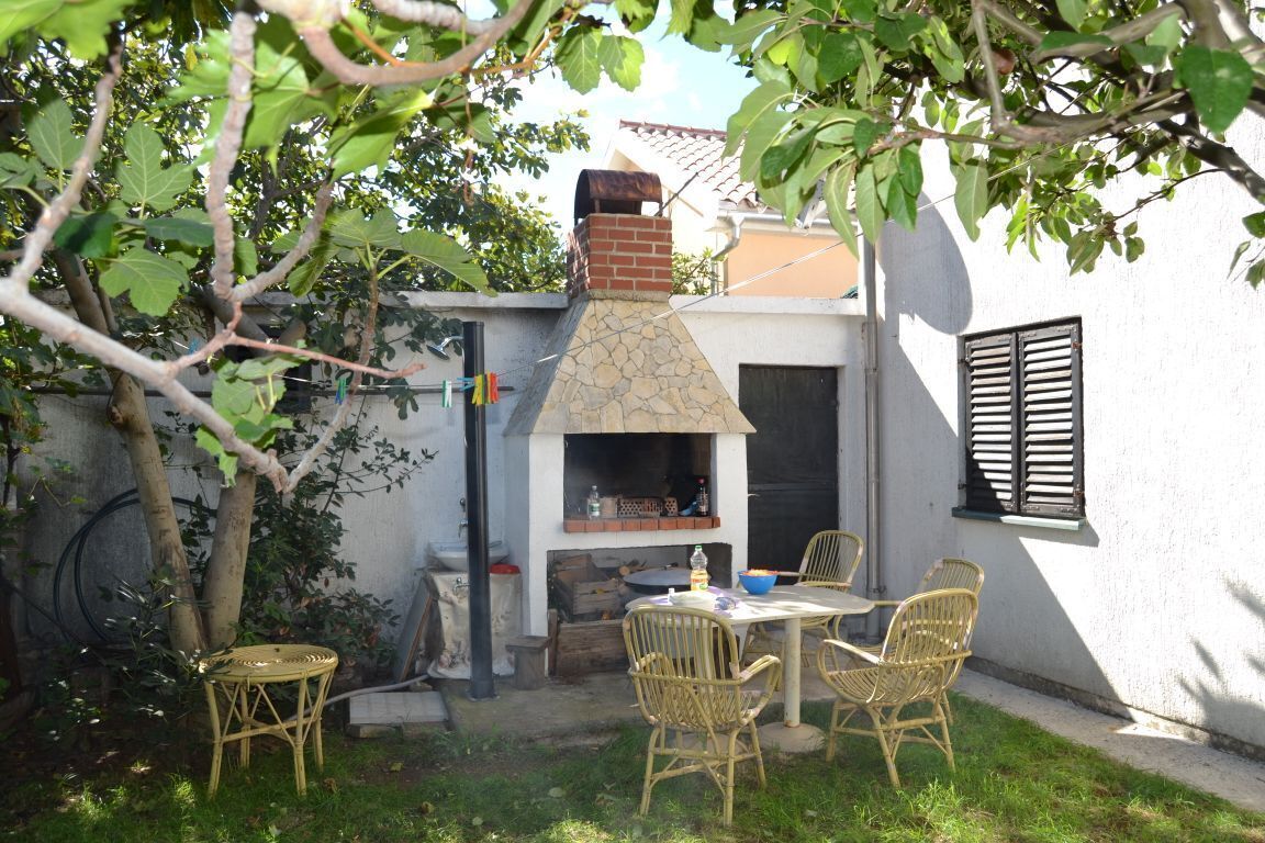 A patio with wicker chairs and a table, a brick oven, and a white wall with shutters.