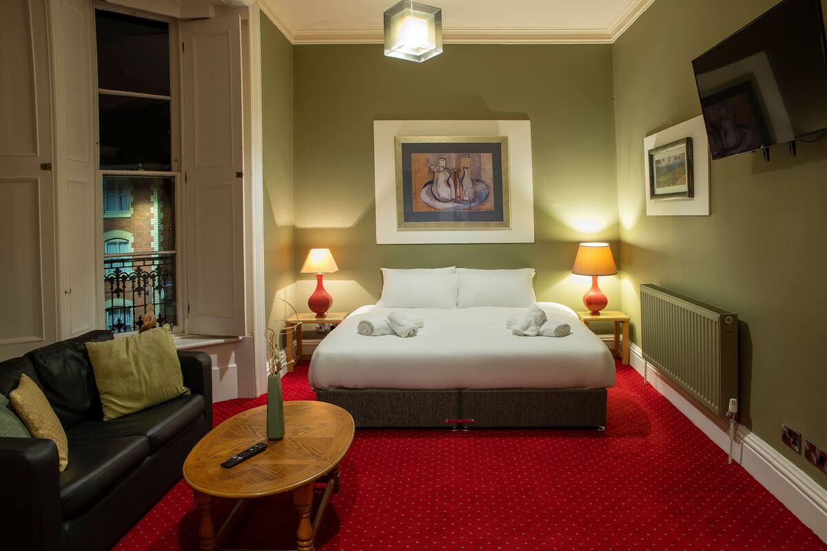 A hotel room with a bed, two bedside lamps, a red carpet, a black leather sofa, and a wooden coffee table.