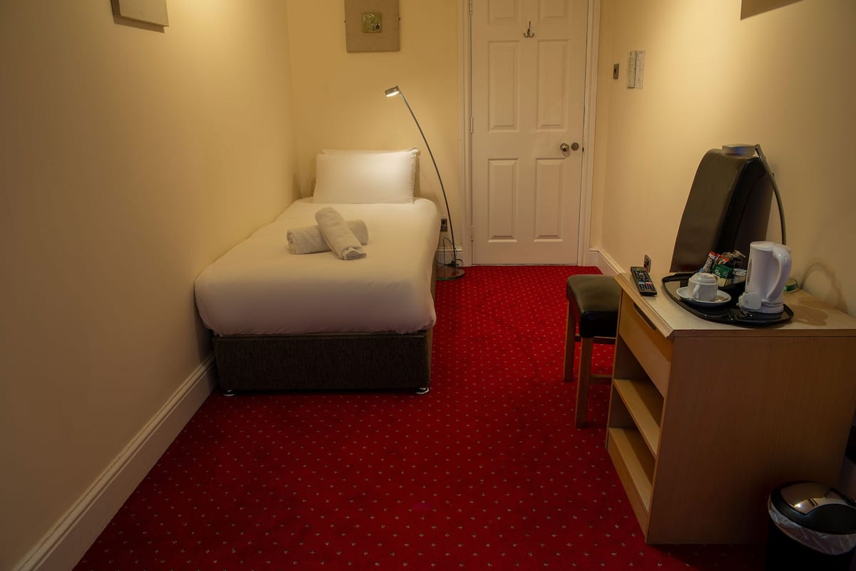 A hotel room with a bed, a desk with a chair, a desk lamp, a coffee maker, and a trash bin.