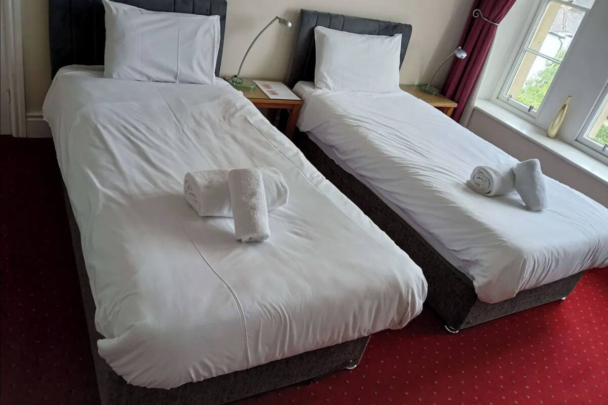 Two single beds with white linens, a nightstand with a lamp, and a window with curtains.