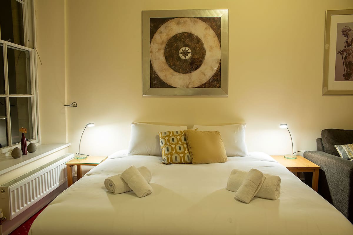 A hotel room with a large bed, two bedside tables, a window, and a painting on the wall.