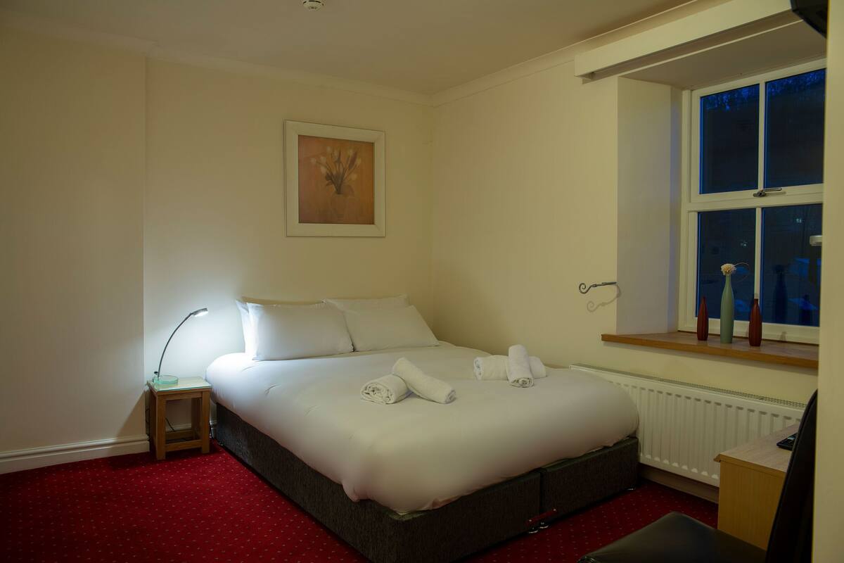 A neatly made bed with white linens and towels, a bedside table with a lamp, a framed picture on the wall, and a window with a view of the outside.
