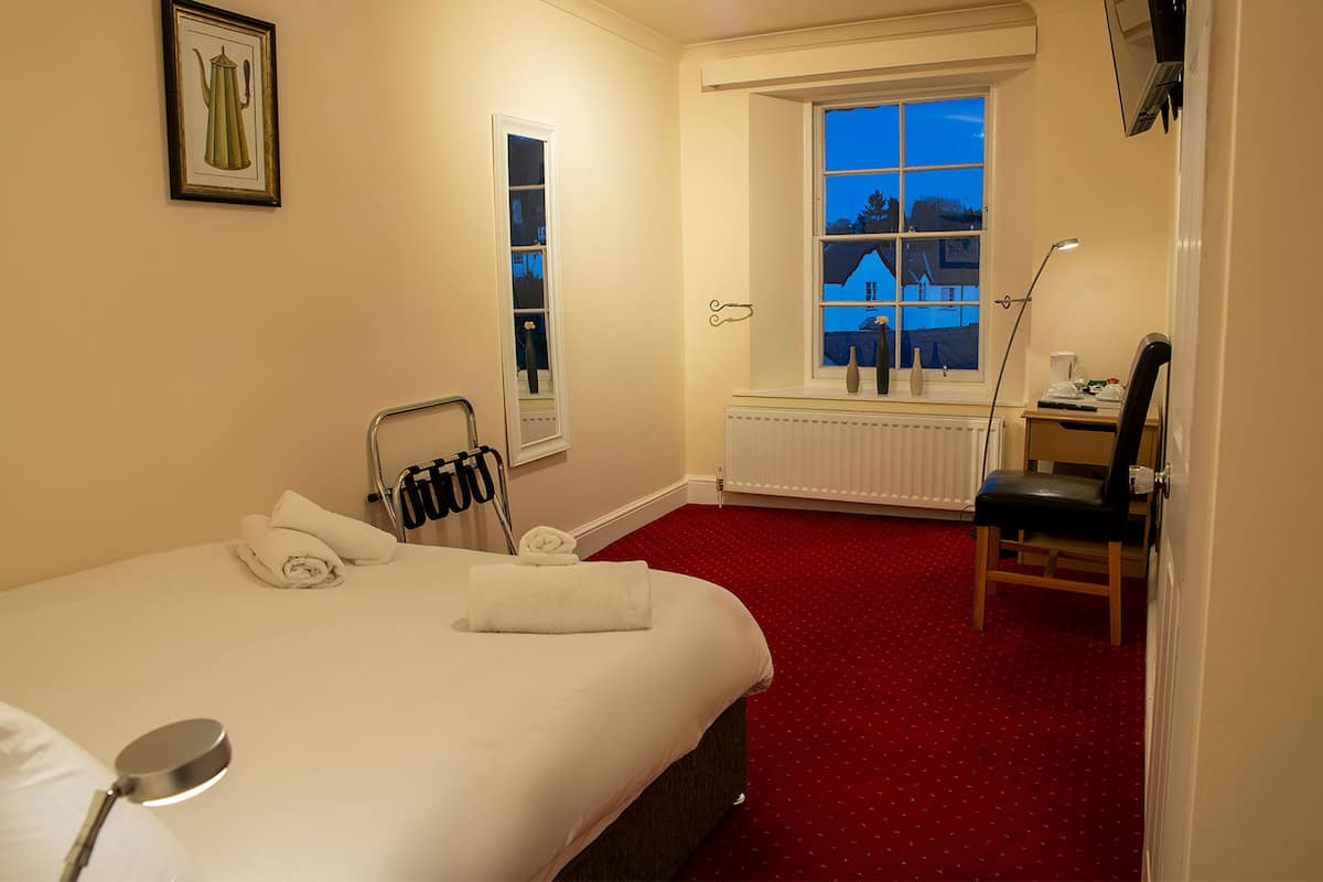 A hotel room with a bed, a desk, a chair, a radiator, a window with a view, and a framed picture on the wall.
