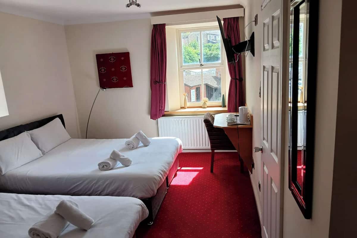 A hotel room with two beds, a desk, a chair, a window with curtains, and a radiator.