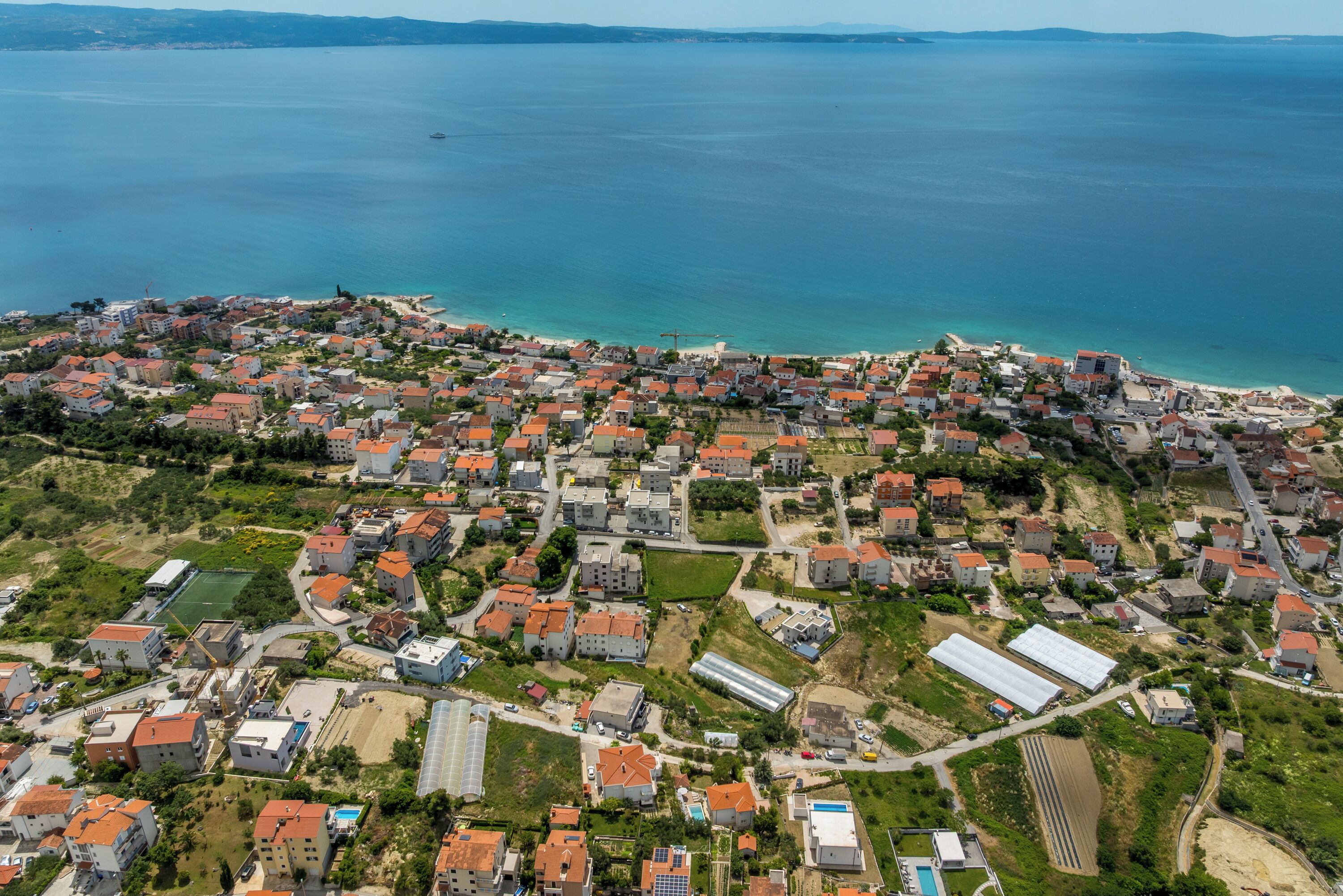 An aerial view of a coastal town with residential buildings, green spaces, and a clear view of the sea.