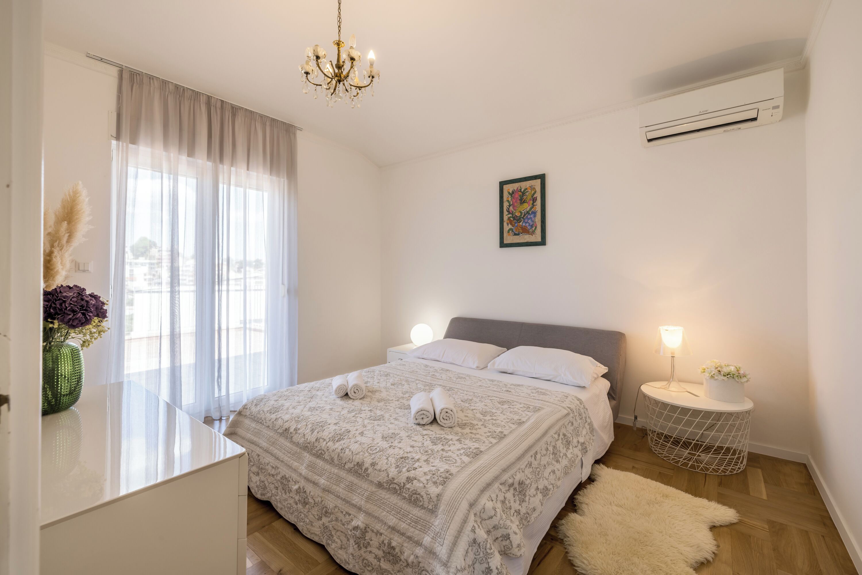 Un dormitorio con cama, lámparas de noche, candelabro, aire acondicionado y ventana con cortinas translúcidas.