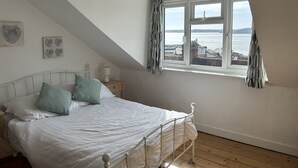 Ein Schlafzimmer mit Bett, Nachttisch, Lampe, einem Fenster mit Vorhängen und Meerblick.