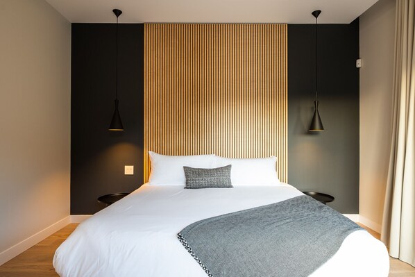 Une chambre moderne avec un grand lit, deux lampes suspendues, un mur avec des lambris en bois et un couvre-lit blanc.
