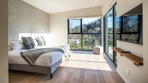 Ein modernes Schlafzimmer mit einem großen Bett, einem Nachttisch und Blick auf einen Wald durch raumhohe Fenster.
