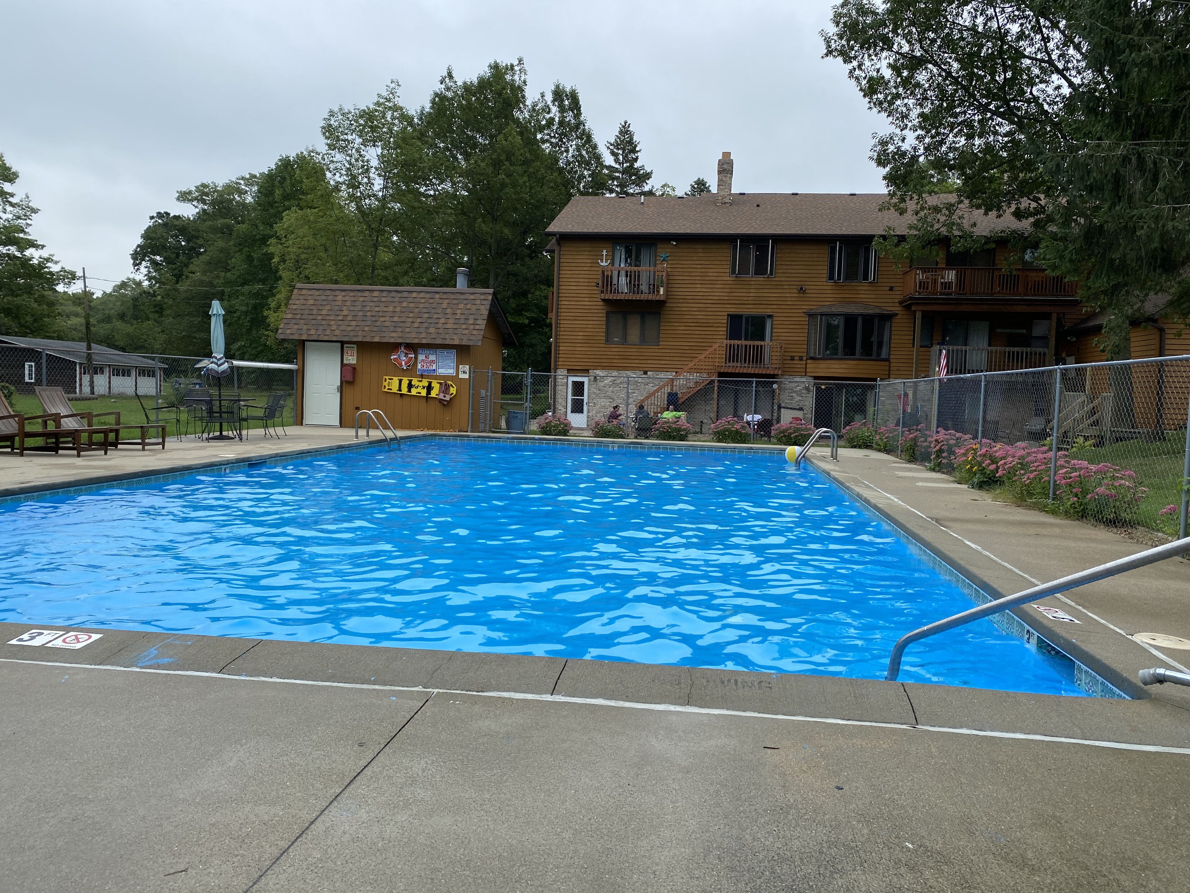 Une piscine avec un poste de surveillance pour les sauveteurs, un petit bâtiment avec une enseigne, et un immeuble à plusieurs étages avec des balcons en arrière-plan.