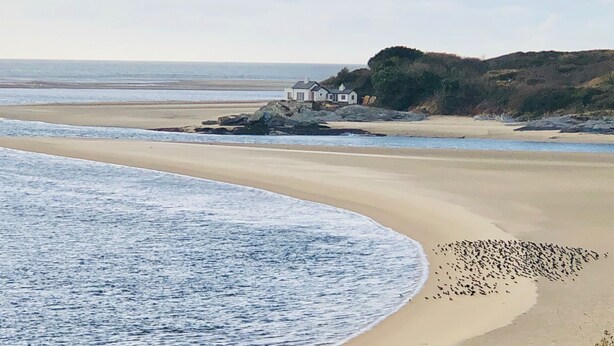 Eine Küstenlandschaft mit Sandstrand, einem kleinen weißen Gebäude auf einem Felsen und ruhiger See.