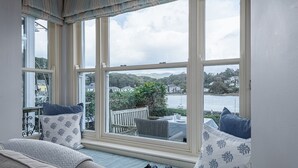 Ein Zimmer mit einem großen Erkerfenster mit Blick auf eine reizvolle Landschaft aus Hügeln und Wasser. Auf einer gepolsterten Bank mit Kissen liegen ein Fernglaspaar.
