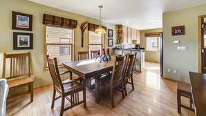 A dining area with a wooden table and chairs, a kitchen in the background, and framed pictures on the walls.