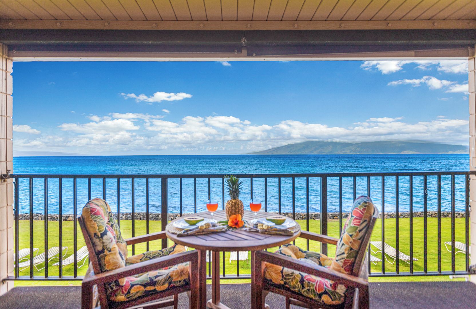 A balcony with a table set for two, overlooking a beach with a clear view of the ocean and a distant mountain.
