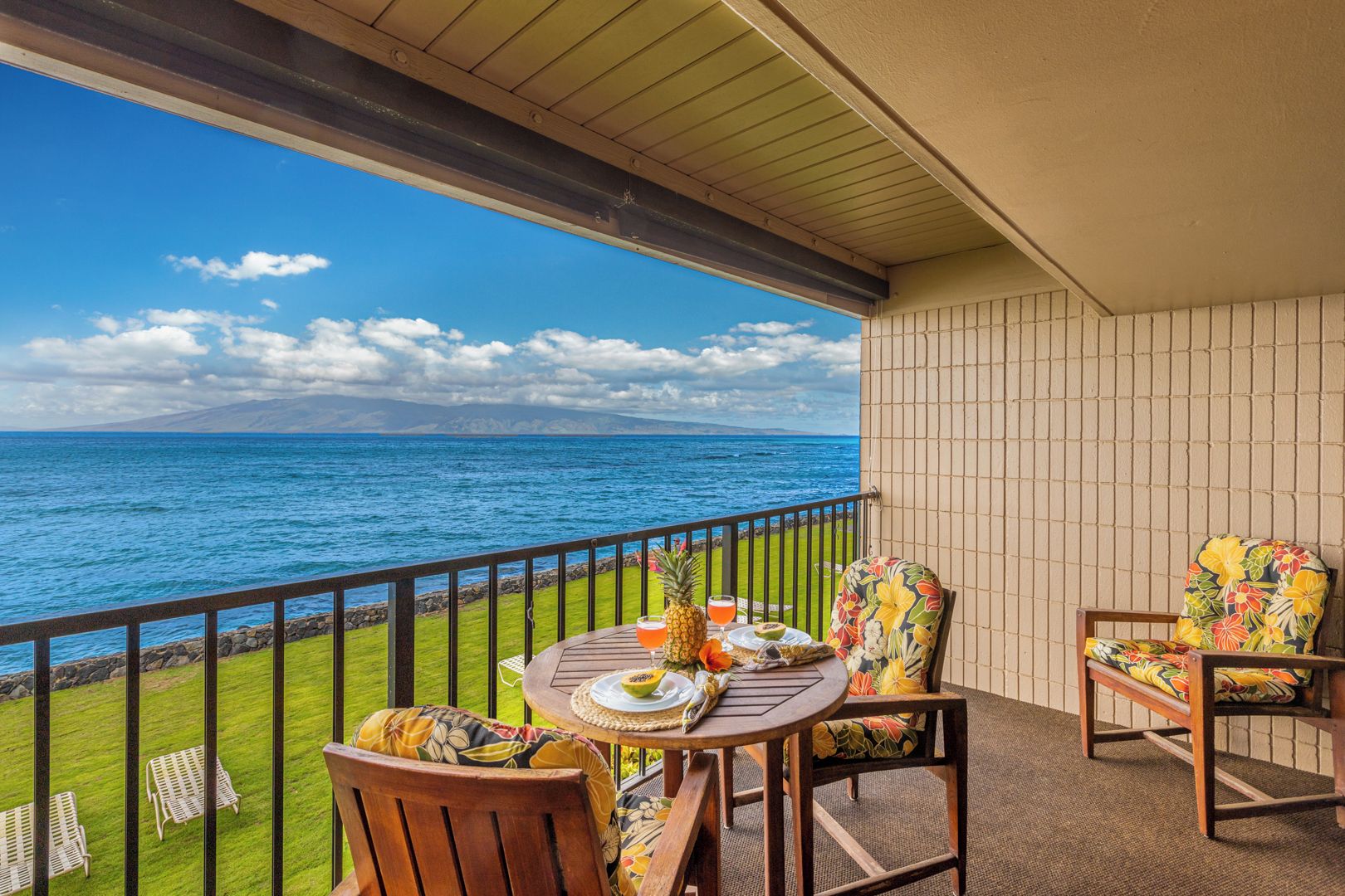 A balcony with a view of the ocean, a table with a drink and fruit, and two chairs with floral cushions.