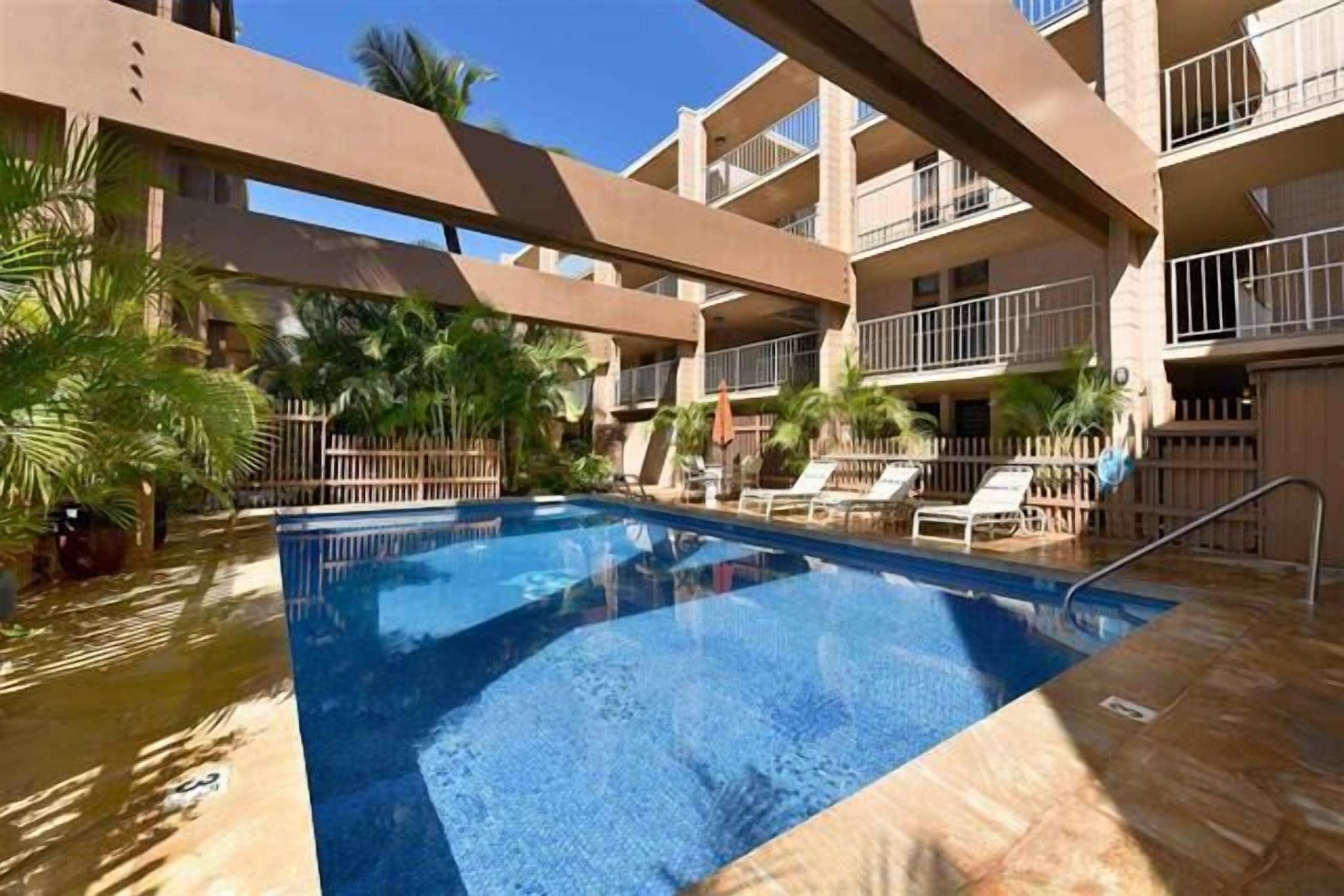 A swimming pool with lounge chairs and palm trees, surrounded by a multi-story building with balconies.