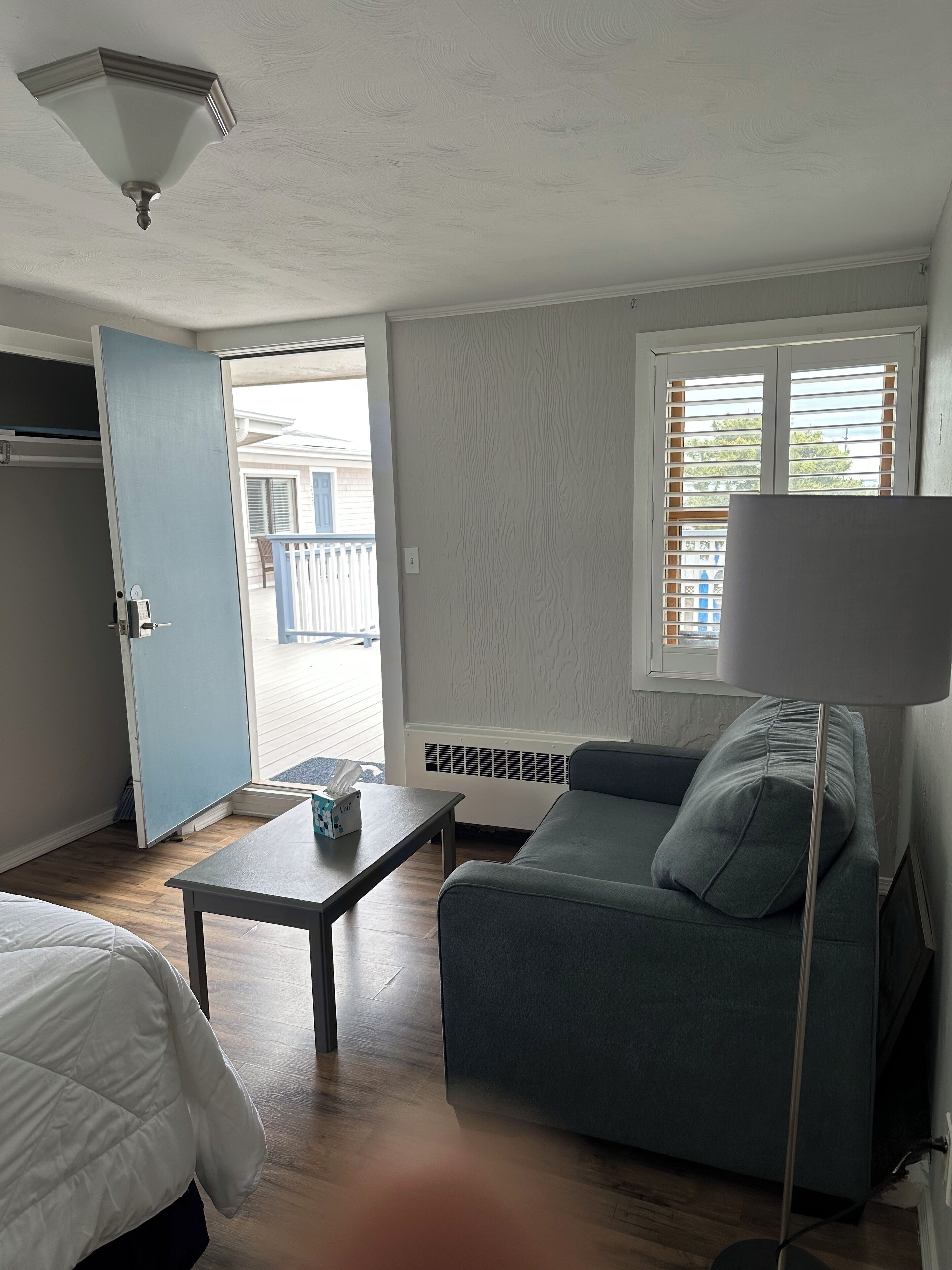 Une pièce comprenant un lit, un canapé, une table avec une boîte de mouchoirs, une lampe et une fenêtre avec des stores.