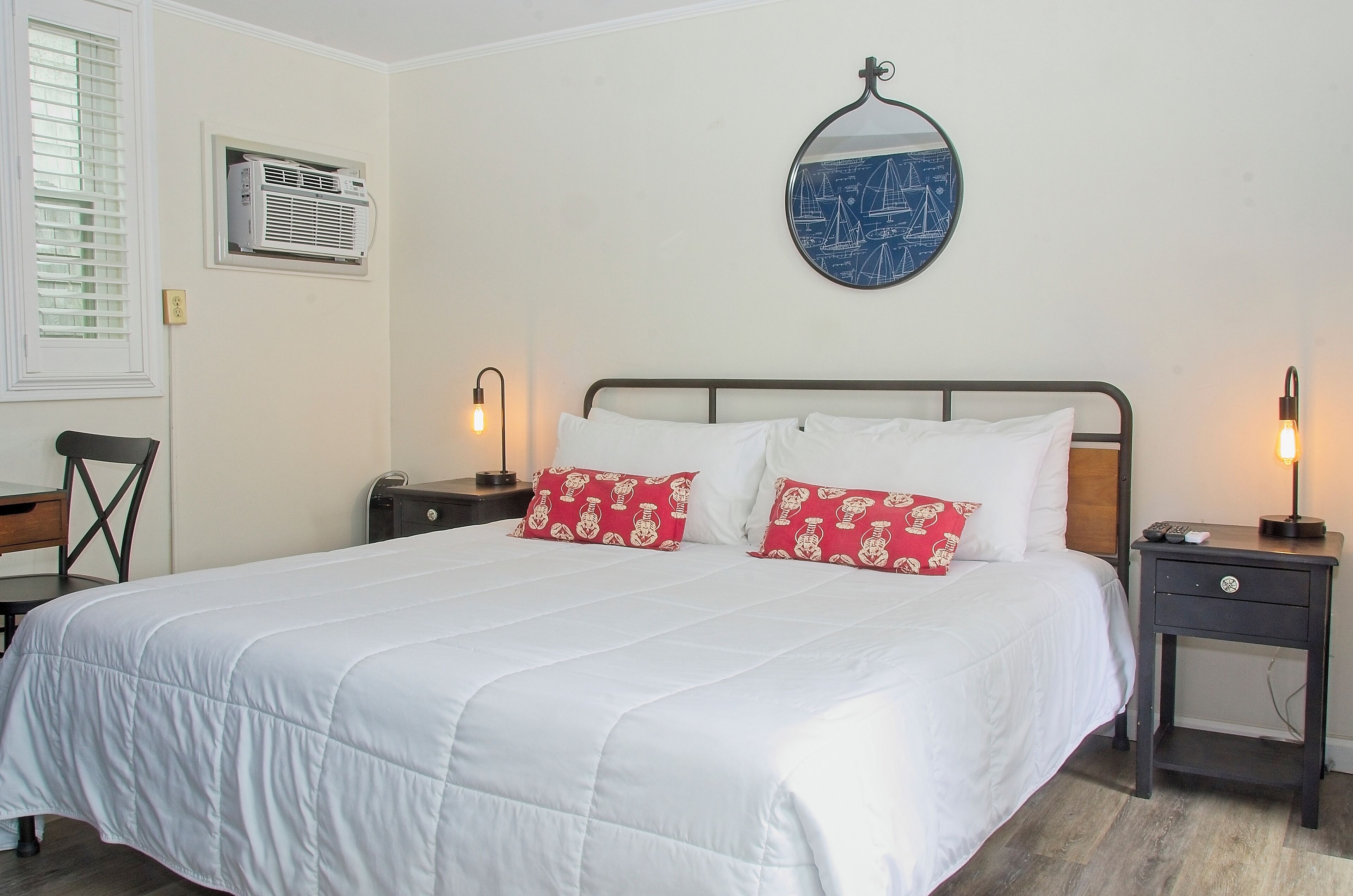 Cama bem arrumada com roupa de cama branca e almofadas vermelhas, mesa de cabeceira com abajur e espelho fixado na parede.