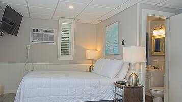 A hotel room with a bed, a desk with a lamp, a TV mounted on the wall, an air conditioning unit, and a bathroom visible through an open door.