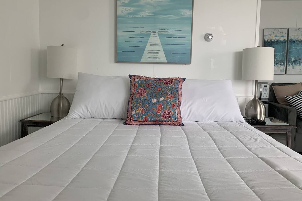 A neatly made bed with a white quilt, a floral pillow, and a painting of a pier on the wall above.