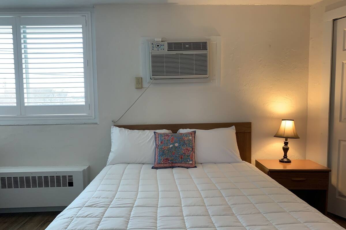 A neatly made bed with a quilted white duvet, a wooden headboard, and a decorative pillow. An air conditioning unit is mounted on the wall above the bed, and a bedside lamp is on the nightstand to the right.