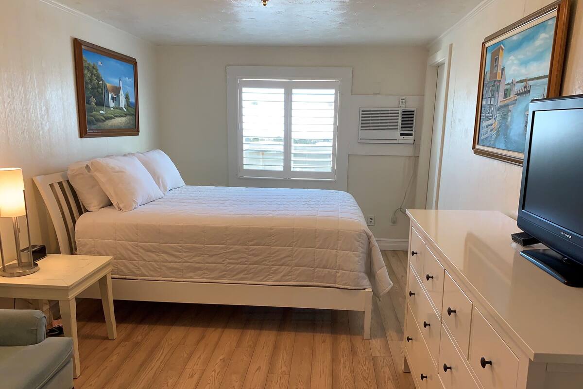 A bedroom with a bed, bedside table, lamp, dresser, TV, and a window with blinds.