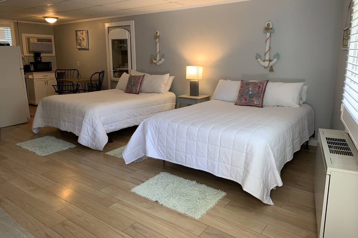 A room with two beds, a desk, a chair, a lamp, and a small kitchenette.