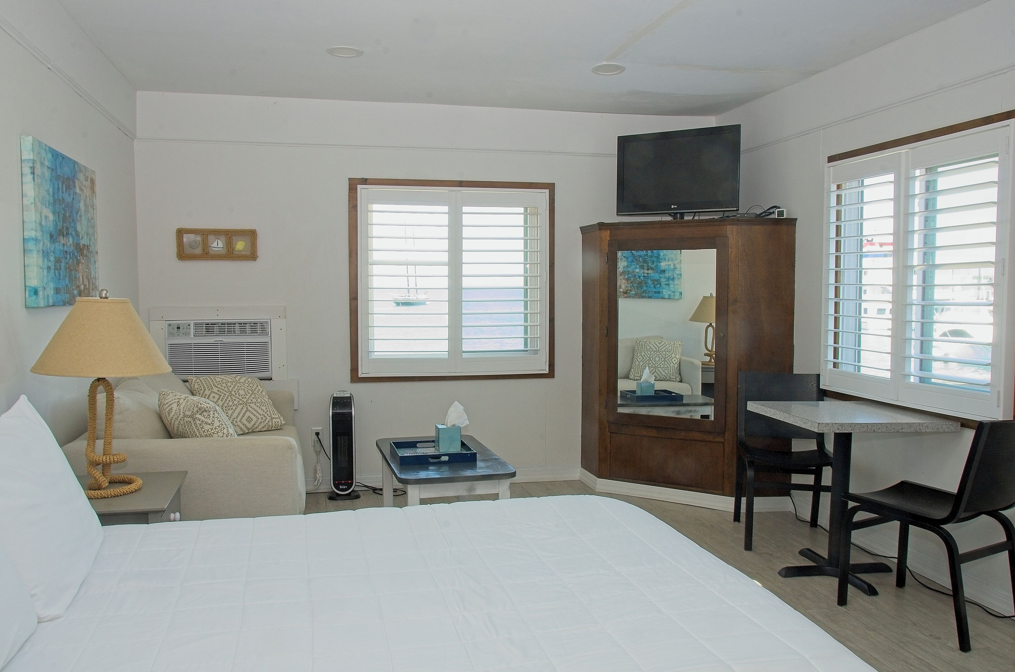 A hotel room with a bed, a desk, a chair, a TV, a window, and a small table with a lamp and tissues.
