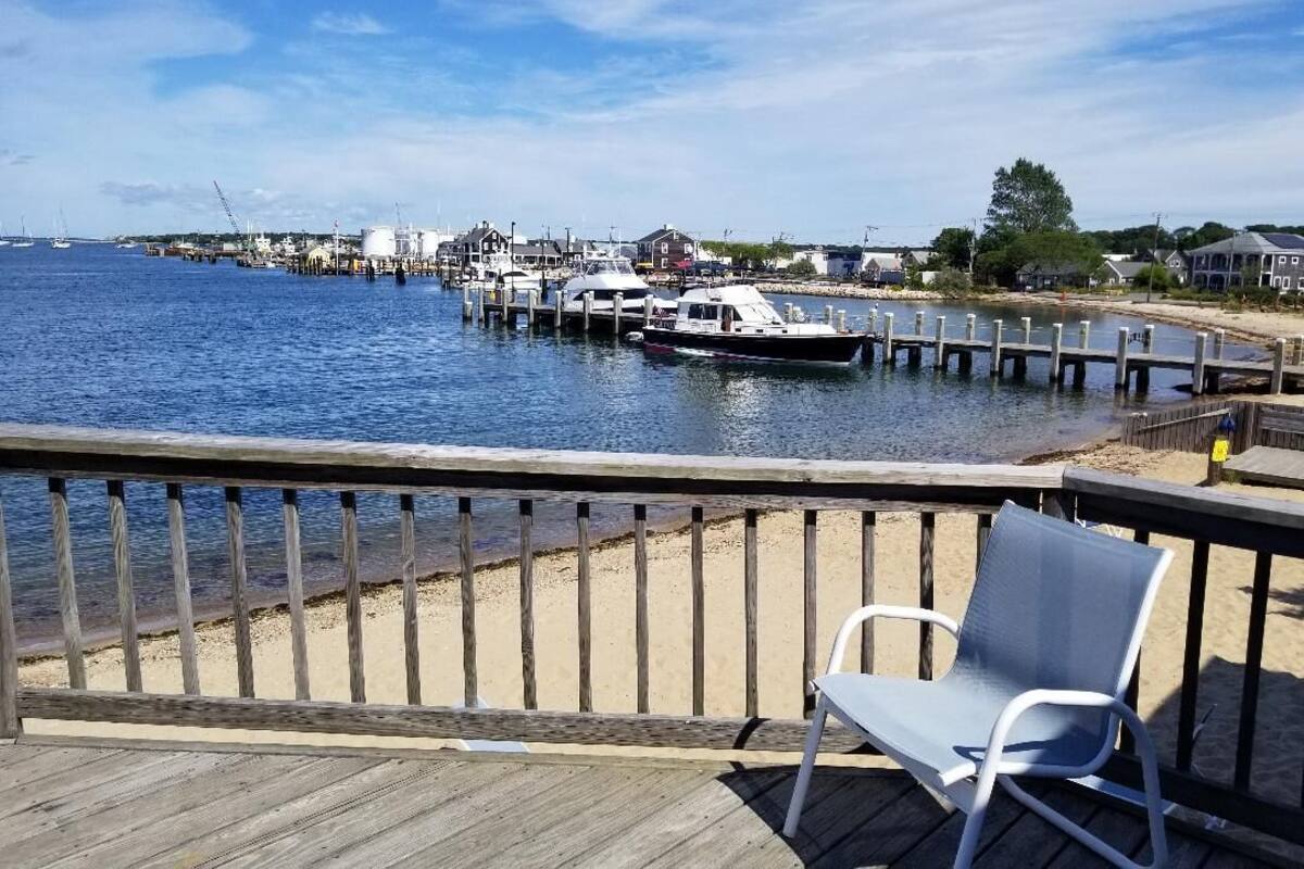 A wooden deck with a metal chair, overlooking a marina with boats docked and houses in the background.