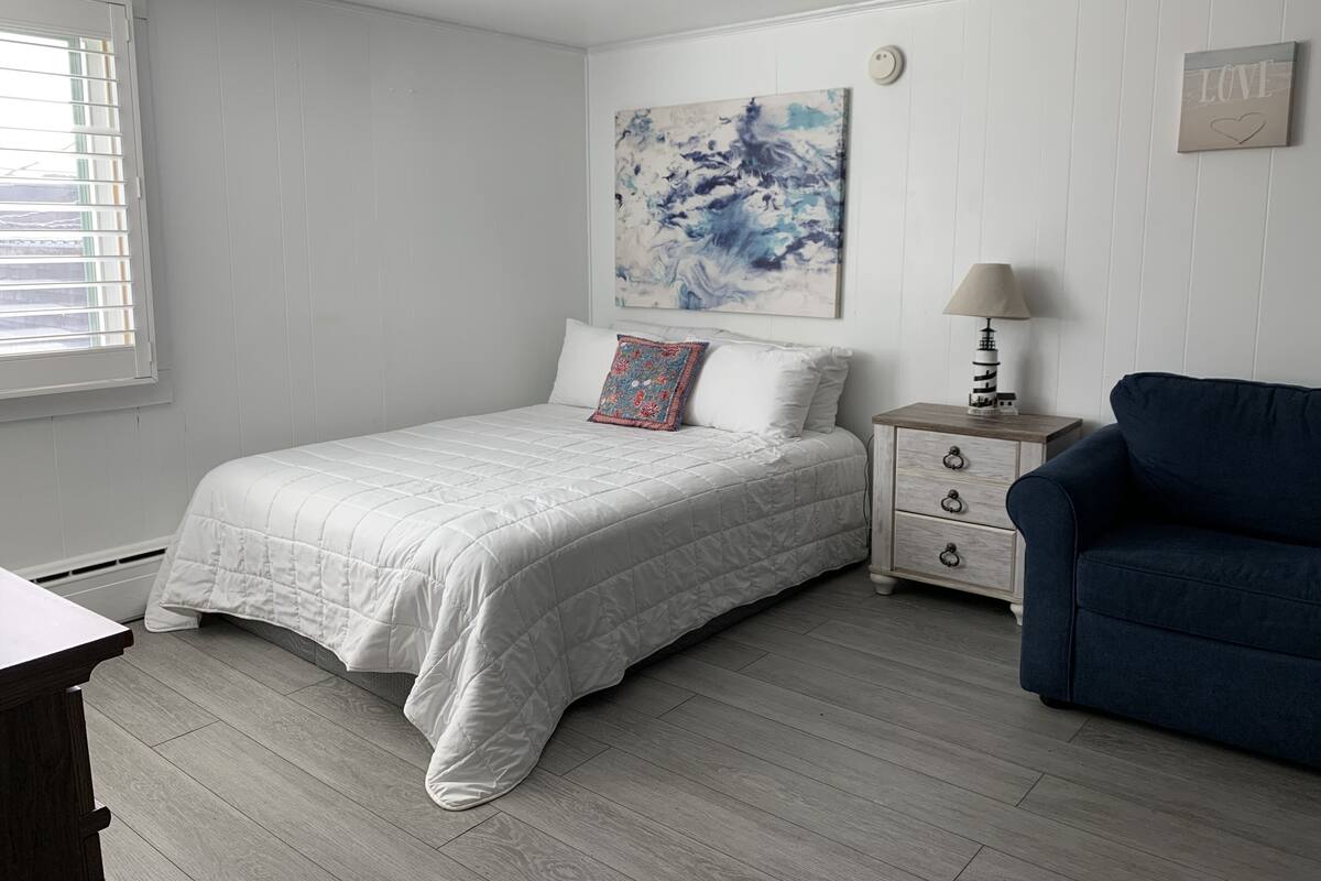 A bedroom with a bed, a nightstand, a lamp, a blue chair, and a painting on the wall.