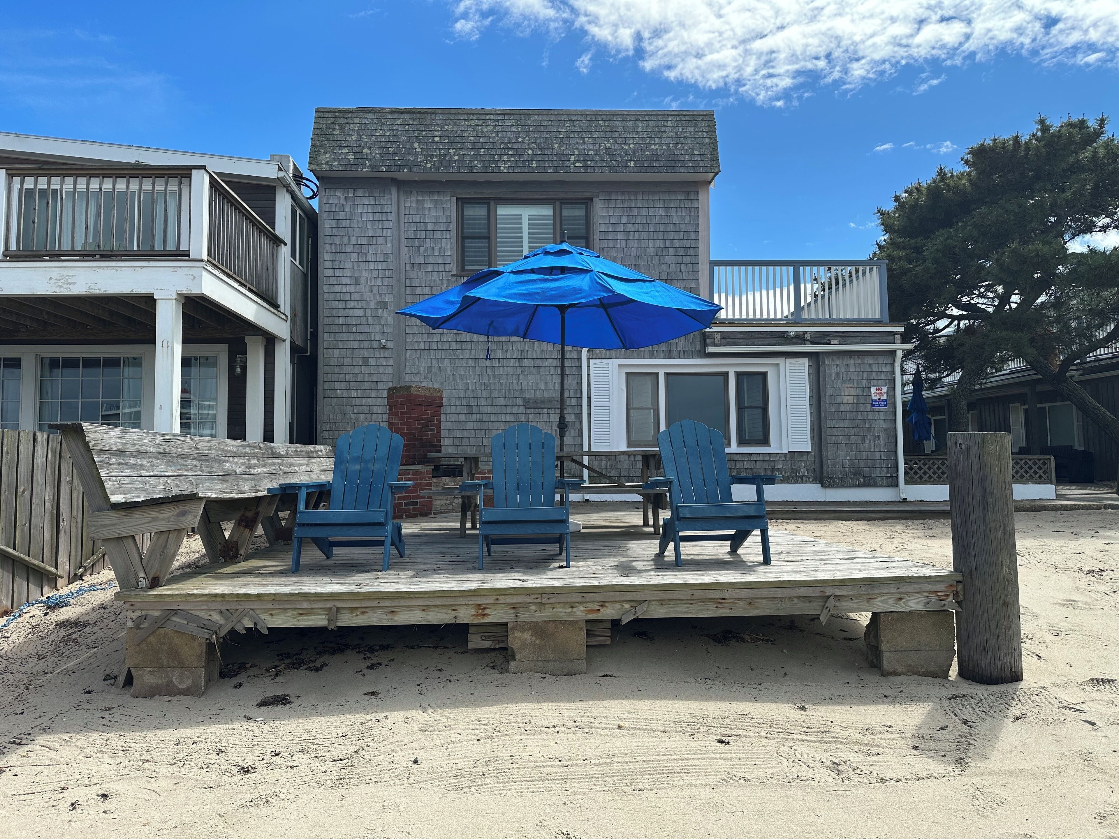 Una terraza de madera con sillas Adirondack azules y una sombrilla azul, con vista a una playa arenosa y un edificio con una chimenea de ladrillo.