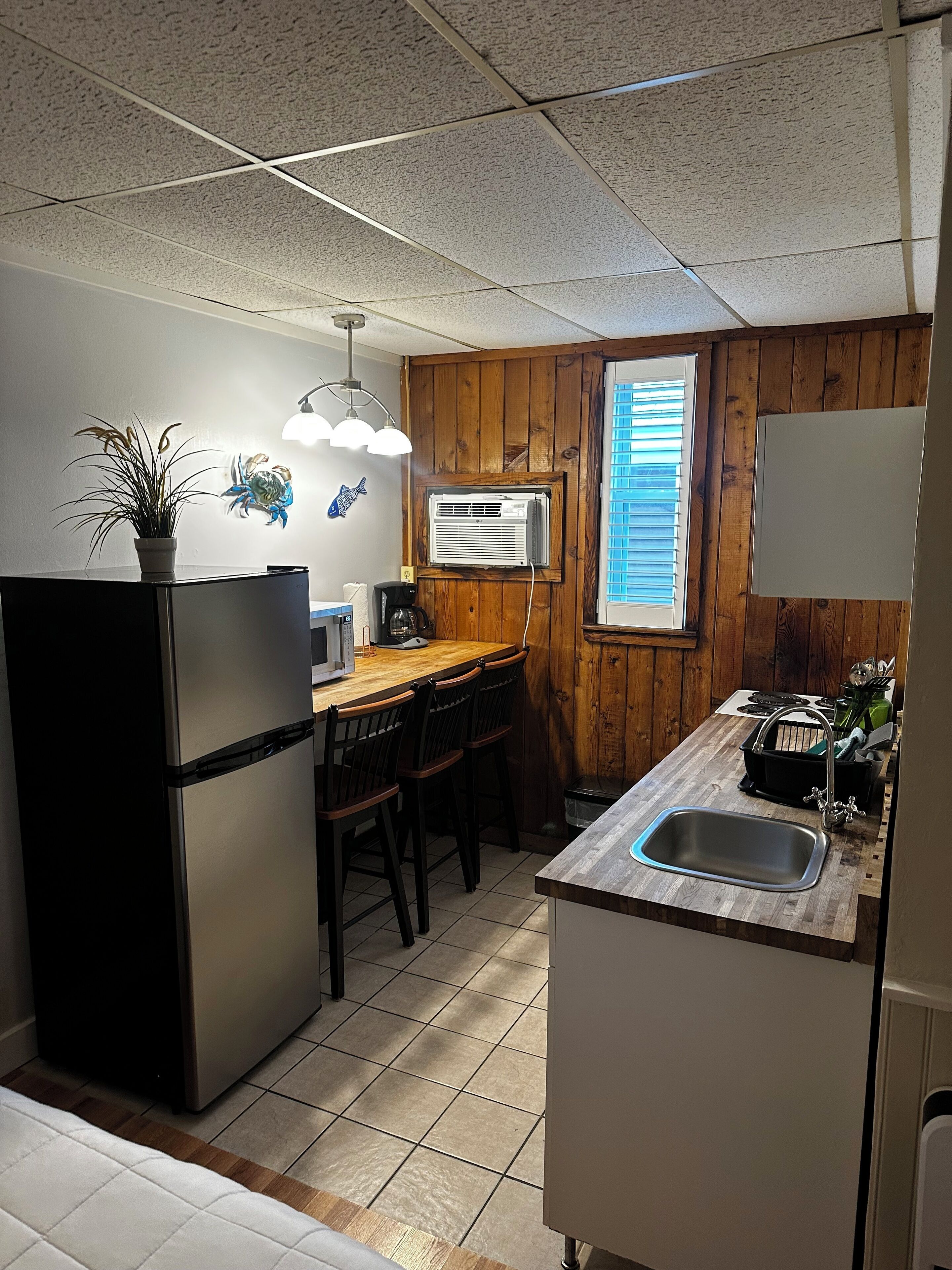 Een kleine keuken met een koelkast, houten wandpanelen, een raam met zonwering, een spoelbak en een eethoek met stoelen.