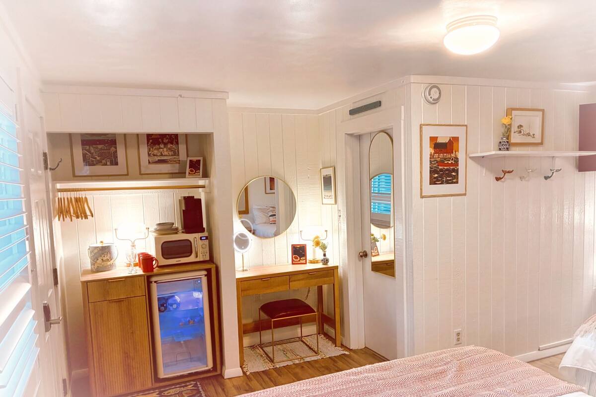 A compact bedroom with a bed, a small desk with a mirror, a microwave, and a refrigerator.