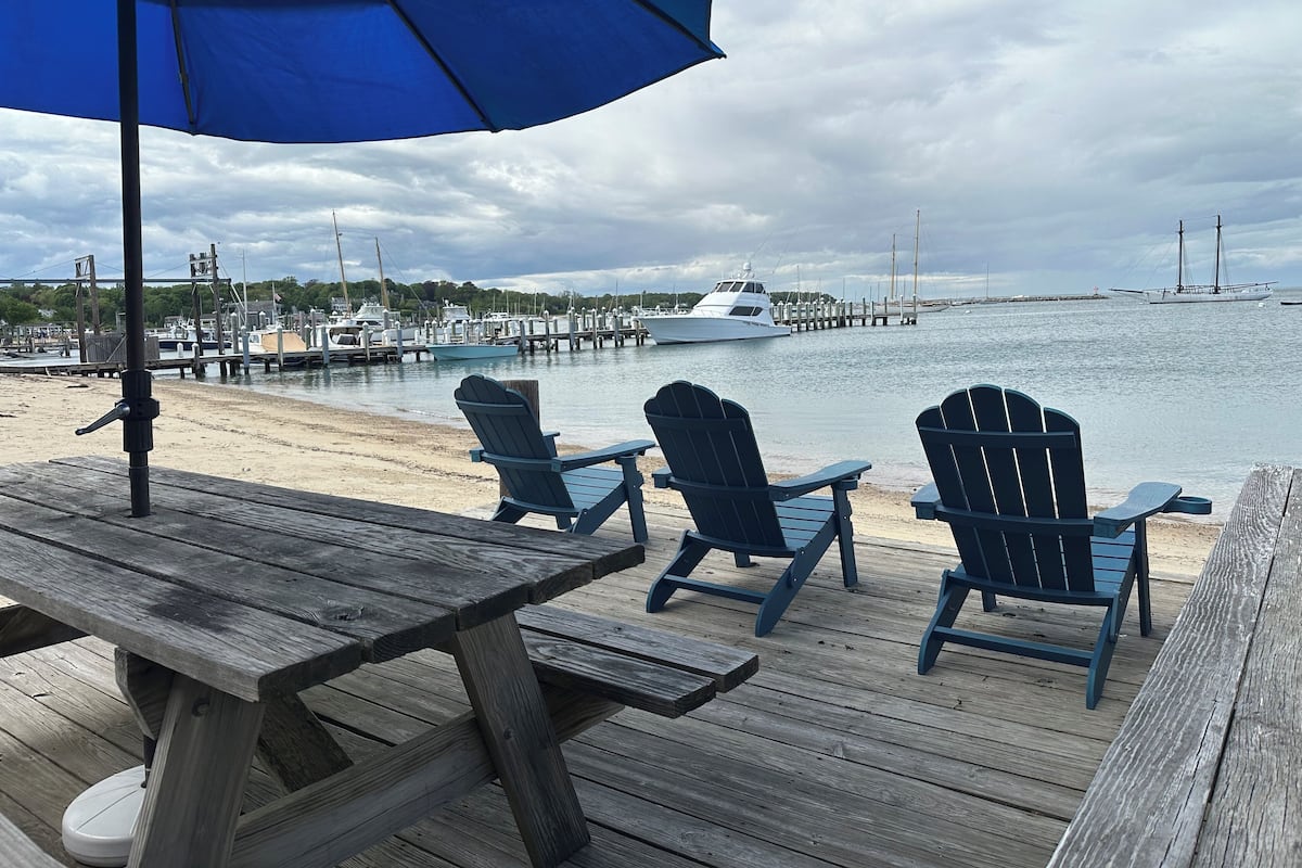 A wooden deck with a picnic table and blue Adirondack chairs overlooking a marina with boats and a dock.