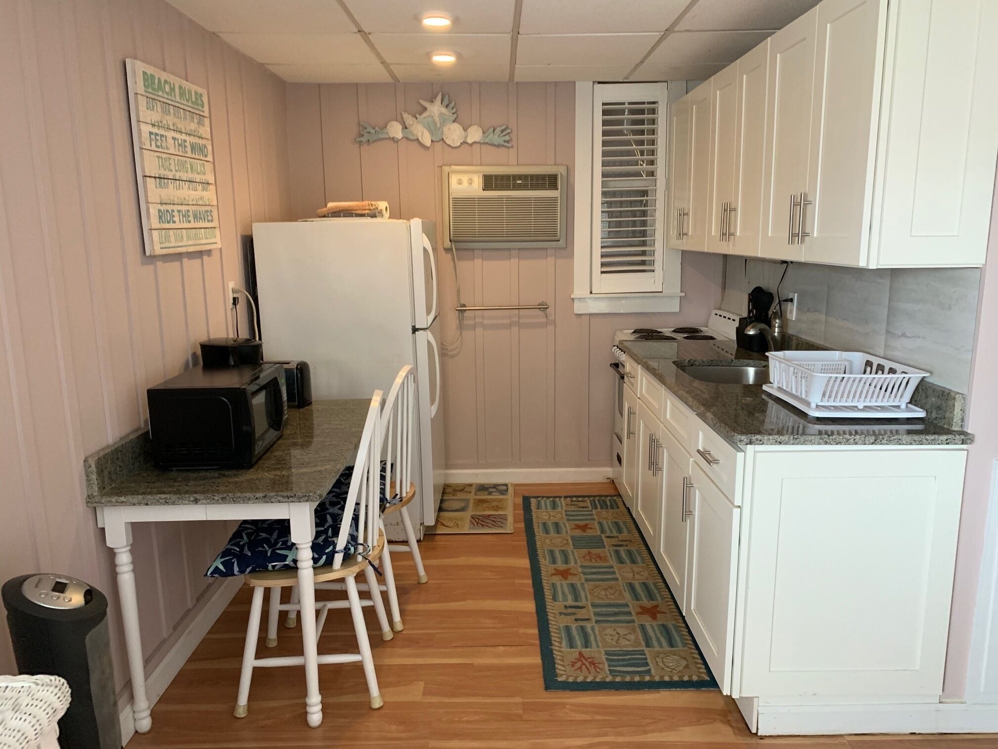 A compact kitchen with a refrigerator, microwave, and stove. There is a small dining area with a chair and a table. The kitchen has white cabinets and a window with blinds.