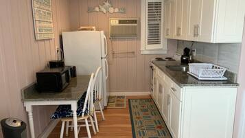 A compact kitchen with a refrigerator, microwave, and stove. There is a small dining area with a chair and a table. The kitchen has white cabinets and a window with blinds.