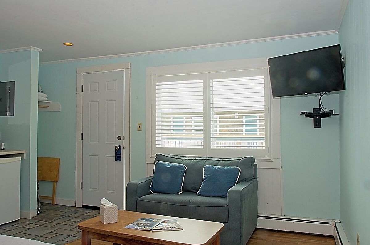A living room with a grey sofa, a coffee table, a television mounted on the wall, and a window with blinds.