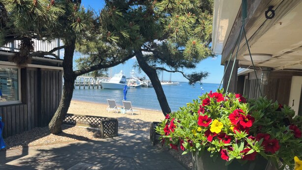 A patio with a wooden table and chairs, a hanging flower pot with red and yellow flowers, and a view of the sea with boats in the background. - Vineyard Harbor Motel - Private Beach (Vineyard Haven)