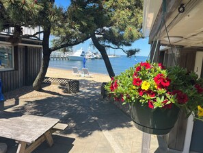 A patio with a wooden table and chairs, a hanging flower pot with red and yellow flowers, and a view of the sea with boats in the background. - Vineyard Harbor Motel - Private Beach (Vineyard Haven)