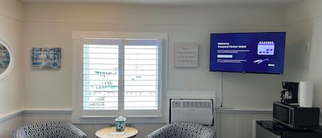 Habitación de hotel con dos sillones estampados, una mesita redonda pequeña con una caja de pañuelos, un televisor mostrando un mensaje de bienvenida, una ventana con persianas y un aire acondicionado instalado en la pared.