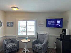 A hotel room with two patterned armchairs, a small round table with a tissue box, a television displaying a welcome message, a window with blinds, and a wall-mounted air conditioner.