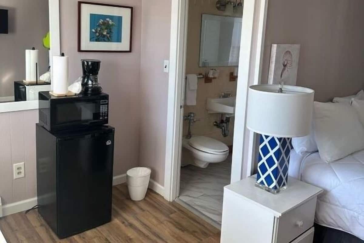 A hotel room with a kitchenette, a bed, a bedside table with a lamp, a bathroom with a toilet and sink, and a door leading to another room.
