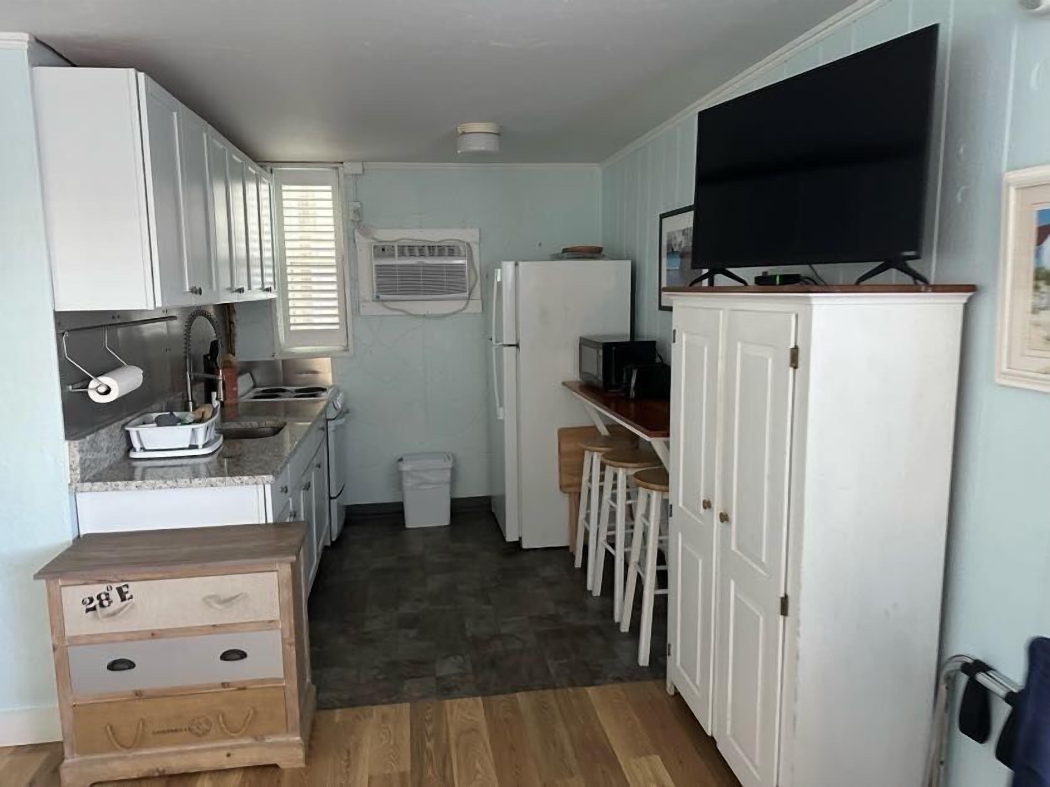 Cocina compacta con armarios blancos, microondas integrado y un refrigerador pequeño. Hay una barra de bar con taburetes y un televisor montado en la pared.