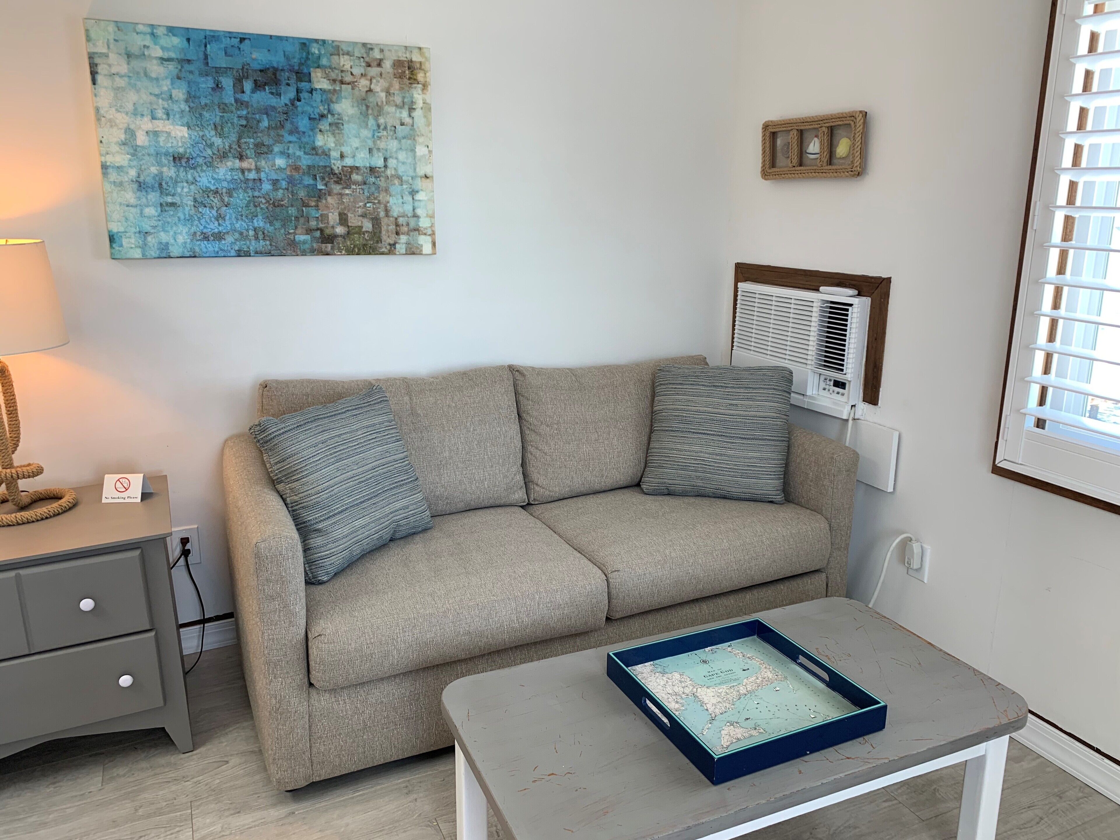 Een woonkamer met een beige bank, een klein tafeltje met een kaart, een lamp en een wandairconditioner.
